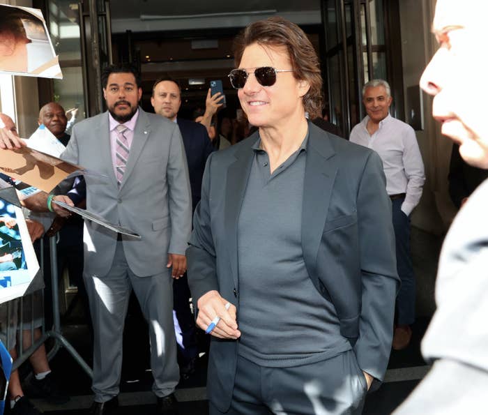 Tom Cruise, in a black suit and sunglasses, smiles and signs autographs for fans outside a building while surrounded by people