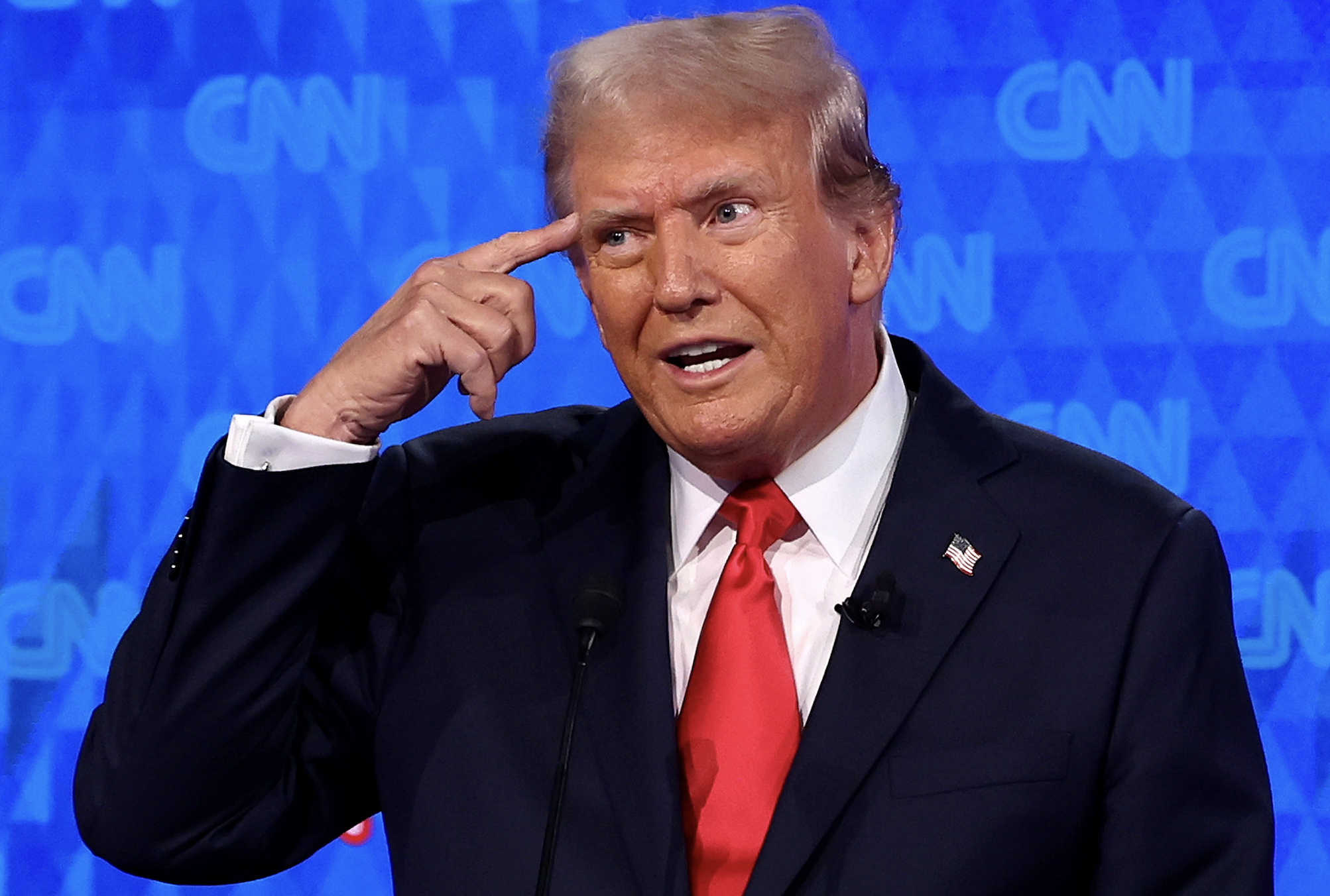 Donald Trump gestures by pointing to his head while speaking at a CNN event. He is wearing a dark suit with a white shirt and a red tie