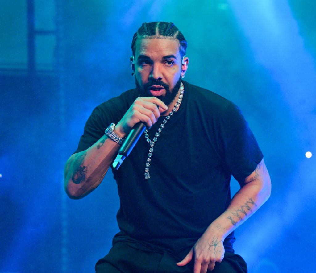 Drake performs on stage, holding a microphone, wearing a black shirt and diamond necklace