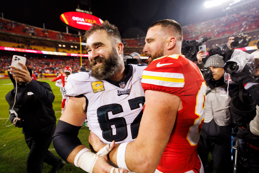Jason Kelce in an Eagles jersey and Travis Kelce in a Chiefs jersey embrace on the football field post-game as photographers capture the moment