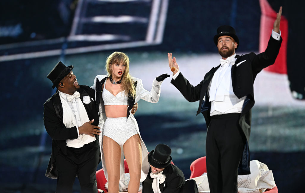 Taylor Swift performing on stage in a white outfit with three male backup dancers dressed in tuxedos and top hats