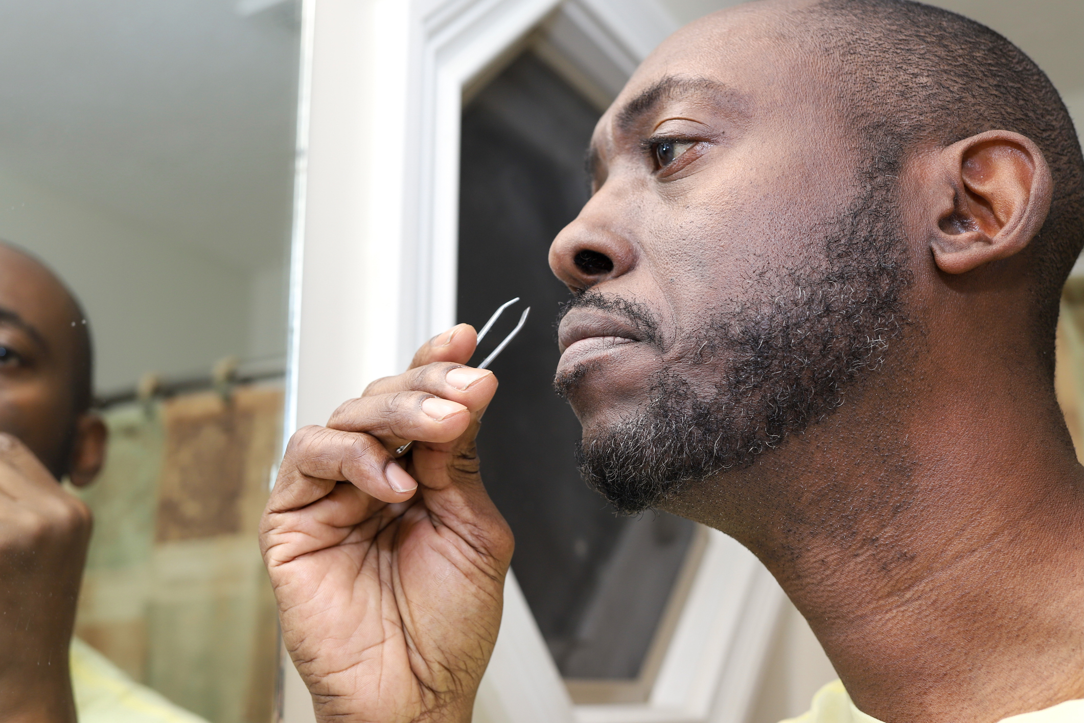 A man stands in front of a bathroom mirror using tweezers to groom his beard