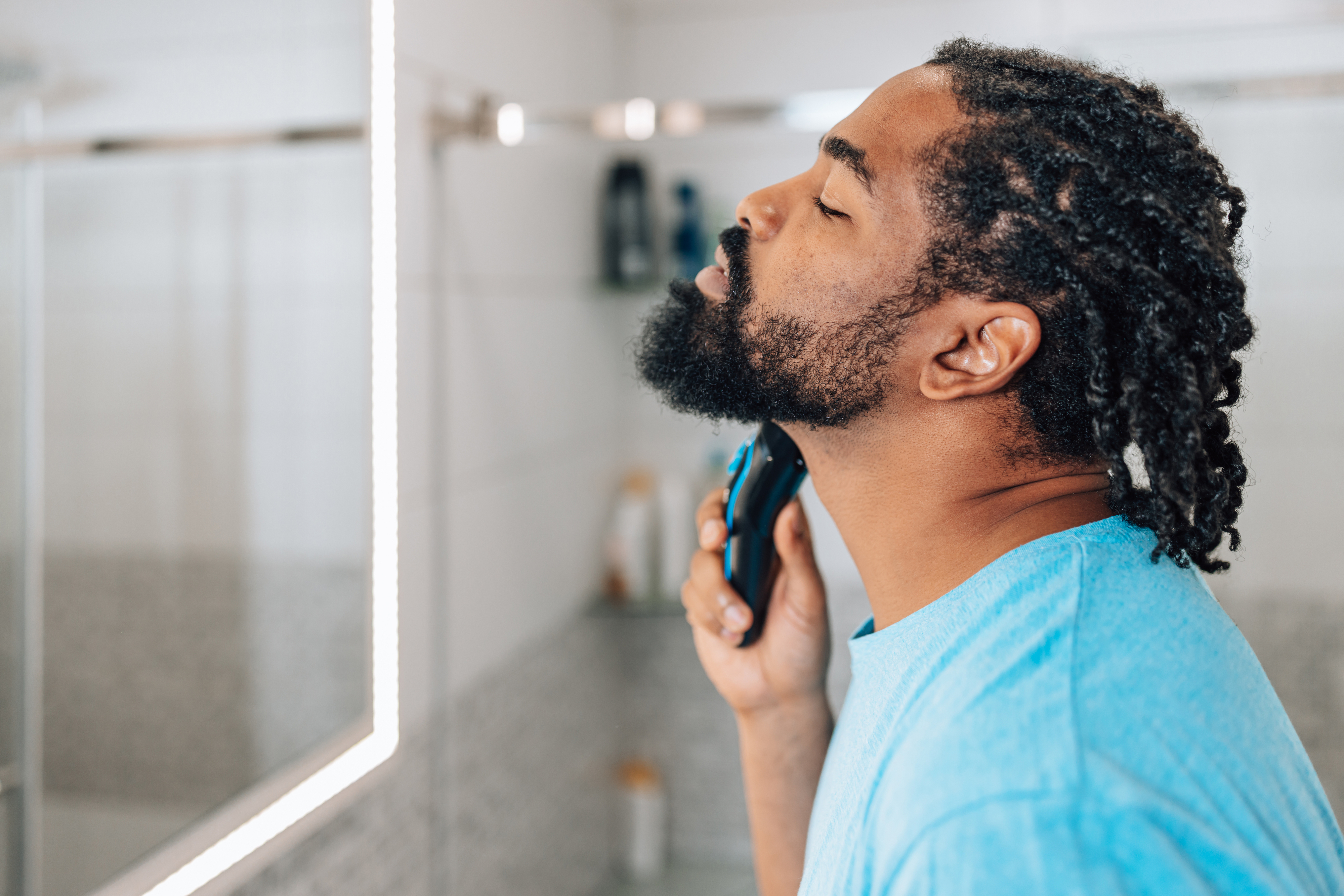 Man in a bathroom grooming his beard with an electric trimmer, wearing a blue shirt