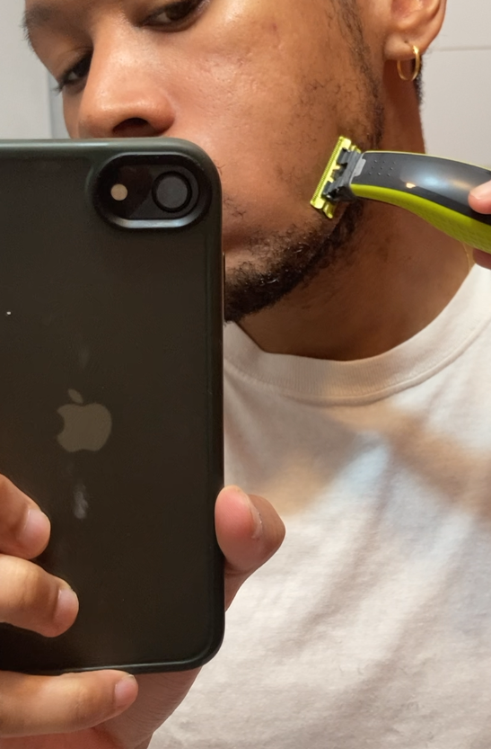 A person in a white shirt holds a phone and shaves their beard with an electric razor