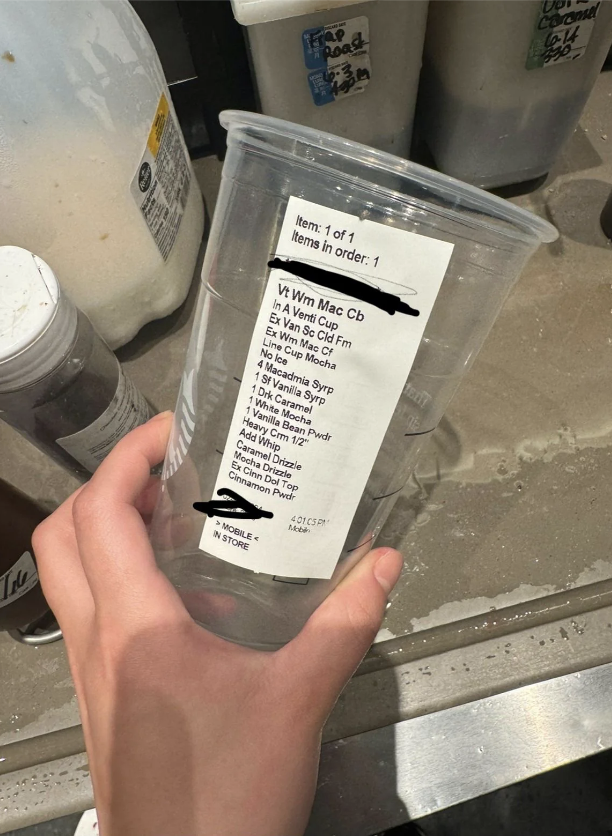 A hand holding a clear plastic cup with a long, detailed coffee order receipt attached to it. Coffee ingredients and instructions are listed