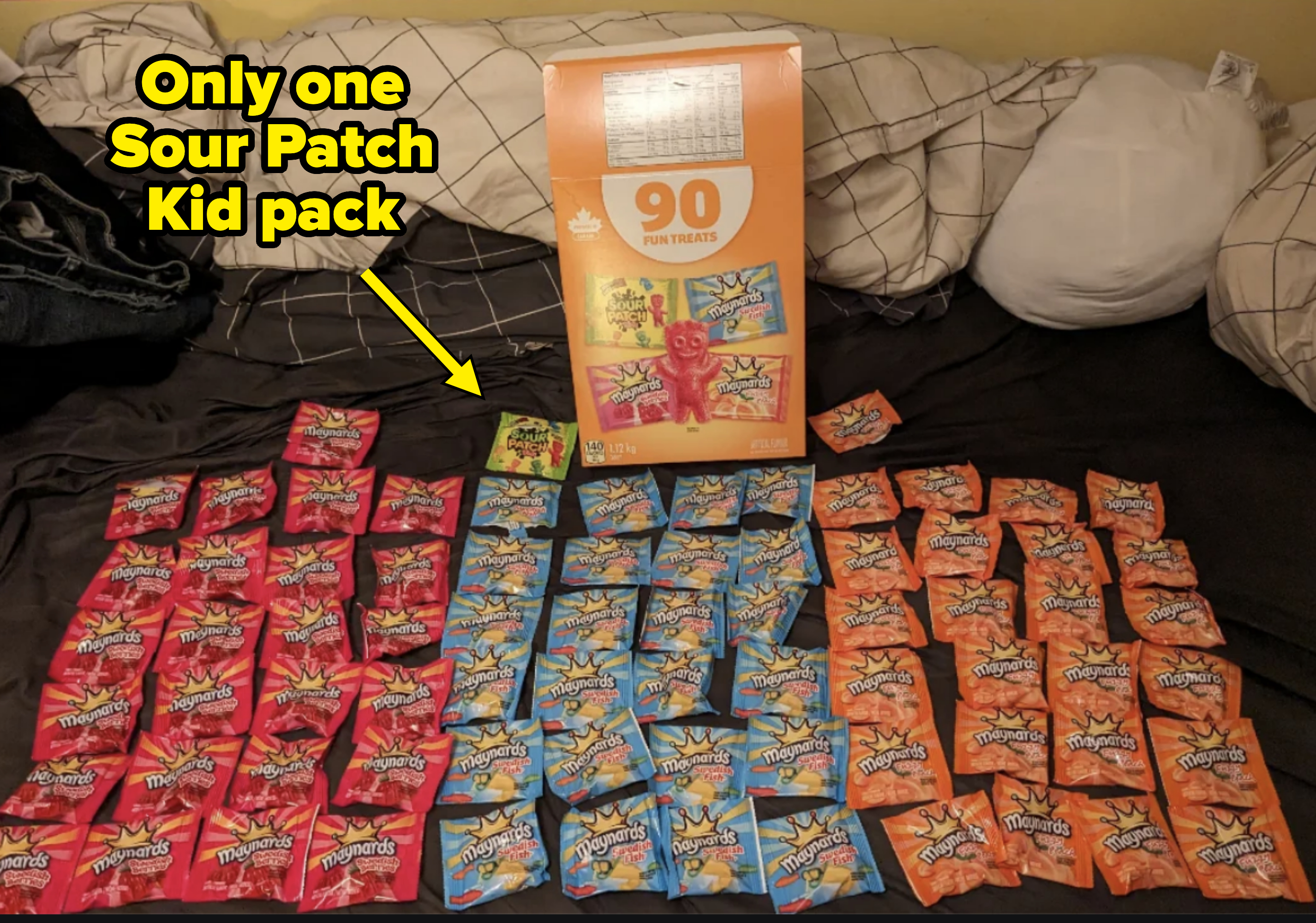 A large box labeled &quot;90 Fun Treats&quot; is surrounded by sorted piles of Sour Patch Kids candy packs on a bed