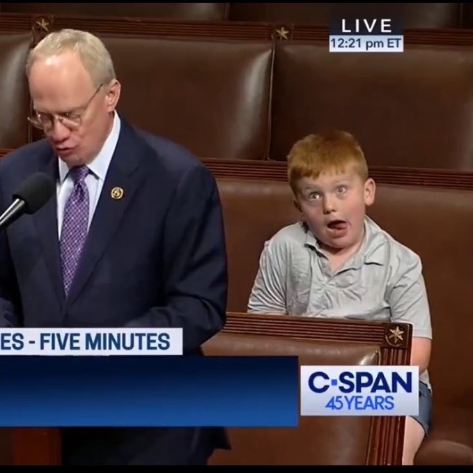 A man in a suit speaks at a podium on C-SPAN. Behind him, a surprised boy with red hair sits in a chair, making a funny face