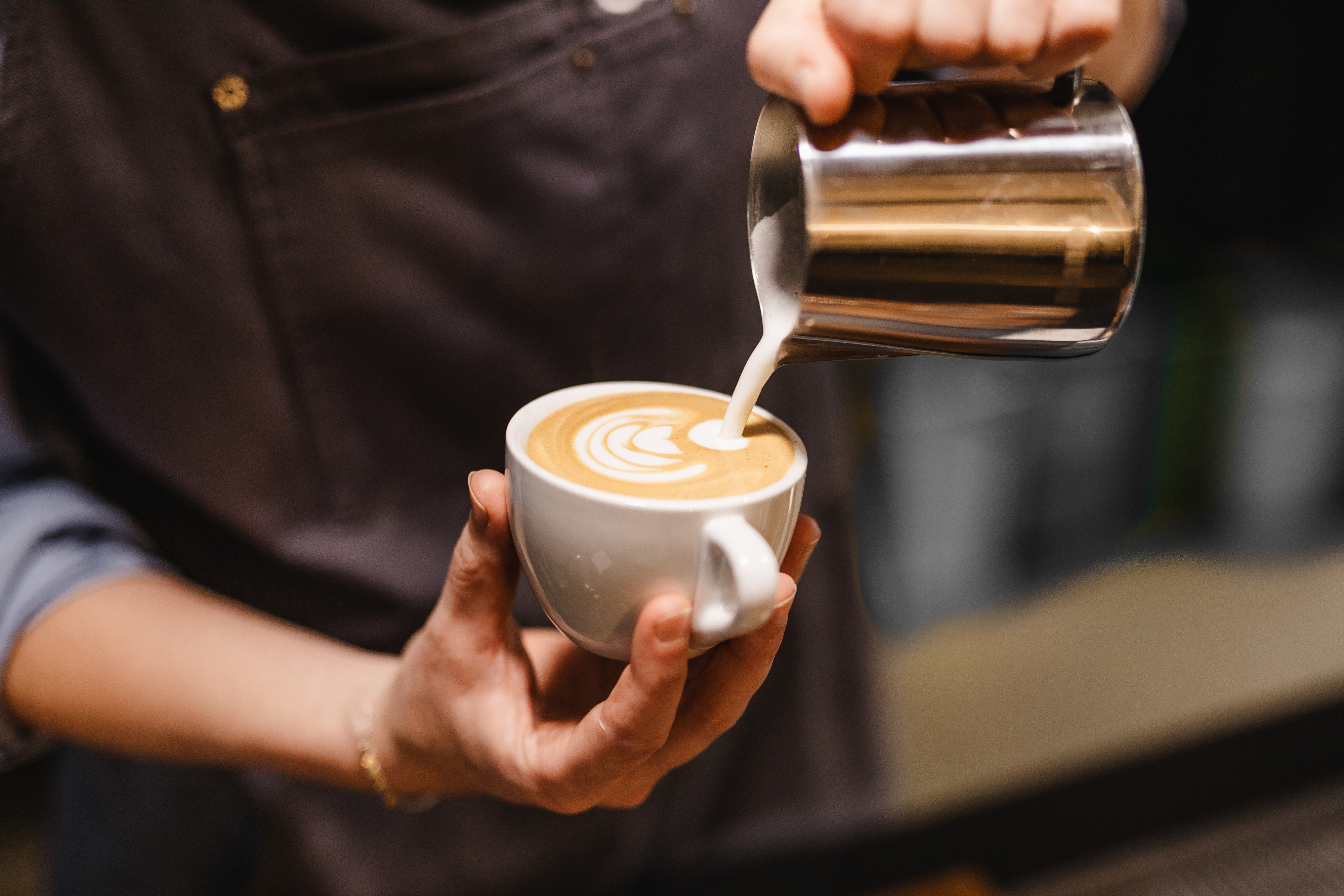 A barista pours steamed milk into a latte, creating a beautiful leaf-shaped latte art design in a white cup