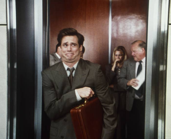 Jim Carrey, in a business suit, awkwardly squeezes into a crowded elevator with a  briefcase in hand. Others in the elevator are also dressed in business attire