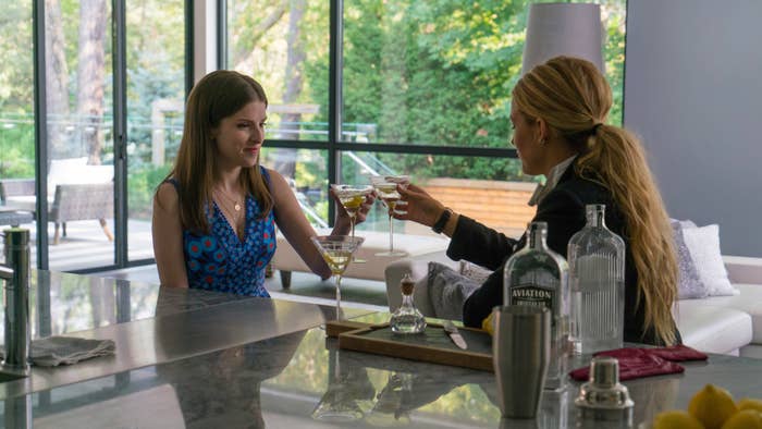 Anna Kendrick and Blake Lively sit at a kitchen counter, toasting with martini glasses. Anna is in a blue patterned dress, and Blake wears a black jacket and white blouse