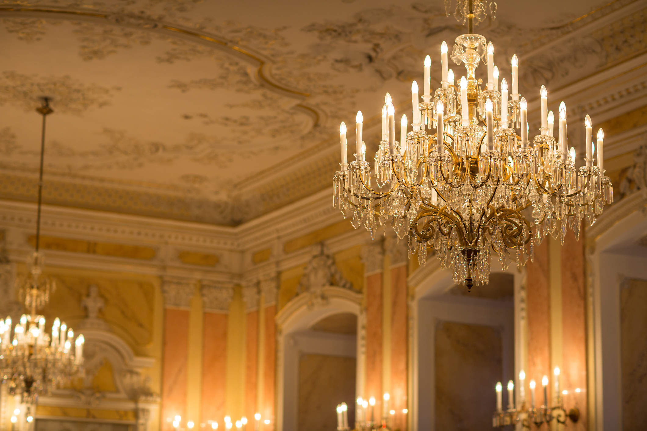 Elegant chandelier hanging in a grand, ornately decorated ballroom with numerous lit candles, high ceilings, intricate wall details, and additional chandeliers in the background