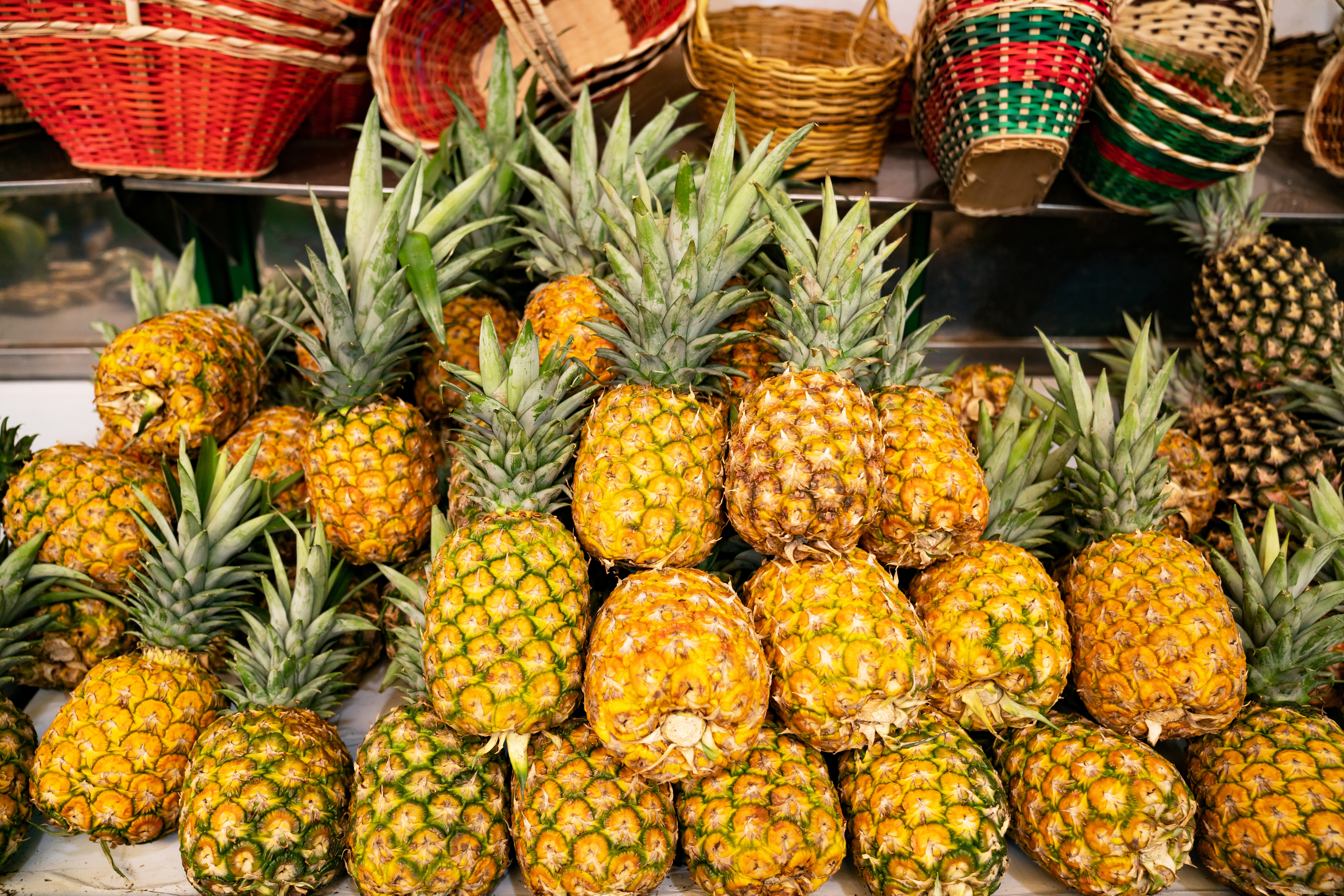 A variety of pineapples are displayed on a market stall. They are stacked in multiple rows with some baskets visible in the background