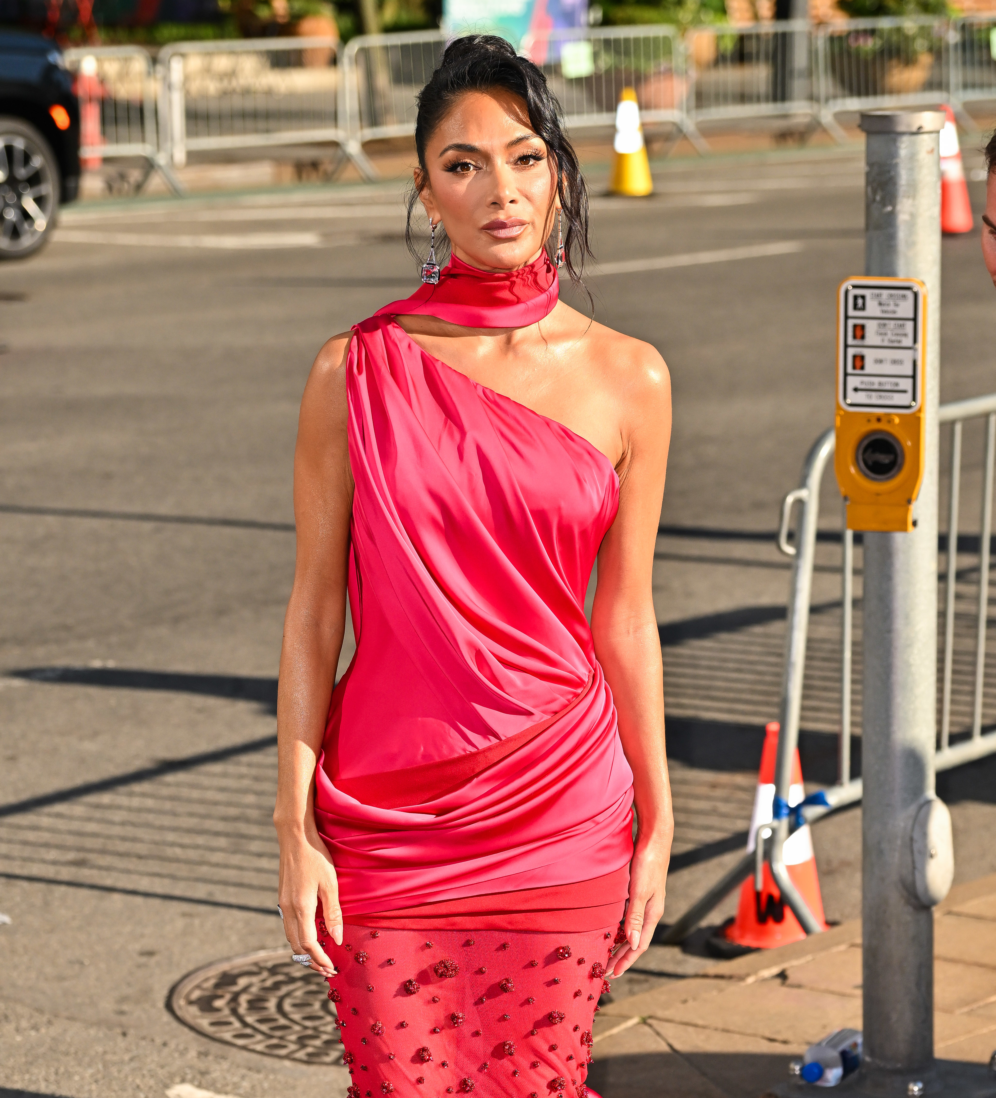 Nicole Scherzinger wearing a one-shoulder, draped red gown with a high neckline and sheer lower skirt, standing outdoors