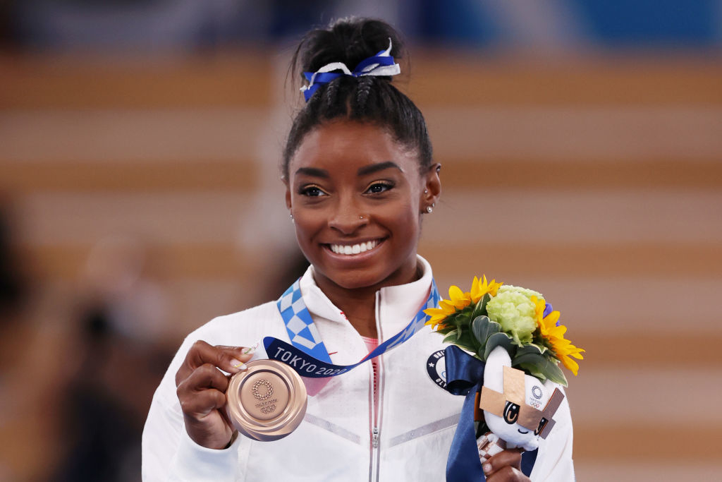 Simone Biles smiles while holding a medal and a bouquet of flowers at the Tokyo 2020 Olympic Games. She is in athletic attire with her hair tied up