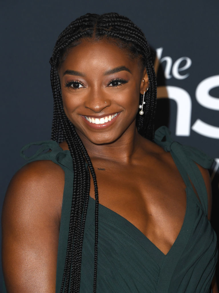 Simone Biles smiling at a red carpet event, wearing a sleeveless dress with braided hair