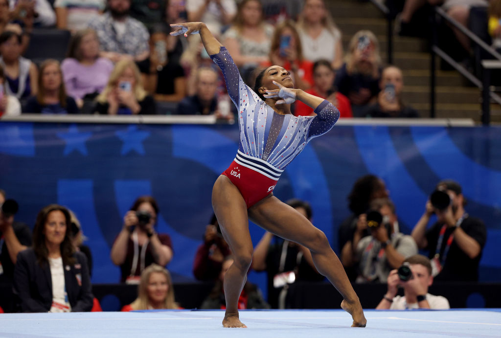Simone Biles performs a gymnastics floor routine at a competition, wearing a leotard with star patterns. Spectators and press watch in the background