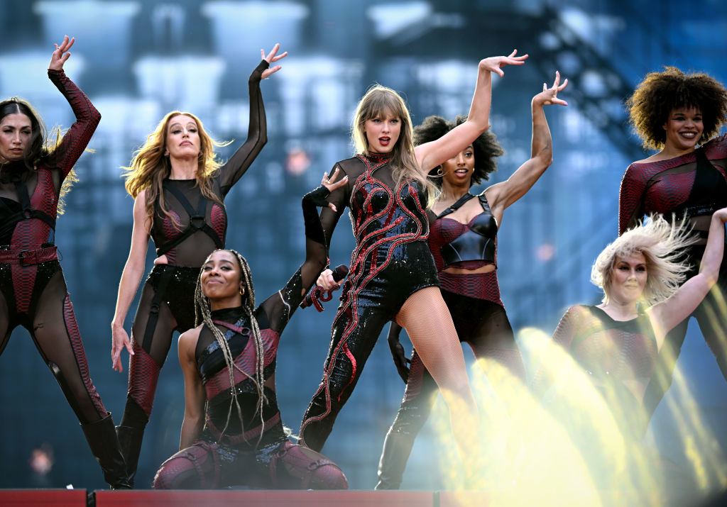 Taylor Swift performs energetically on stage with backup dancers, all dressed in stylish, dark stage outfits