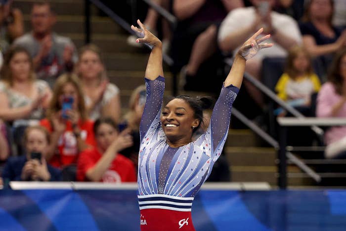 Simone Biles raises her arms in triumph, smiling enthusiastically, wearing a stylish gymnastics leotard with intricate patterns, in front of an applauding crowd