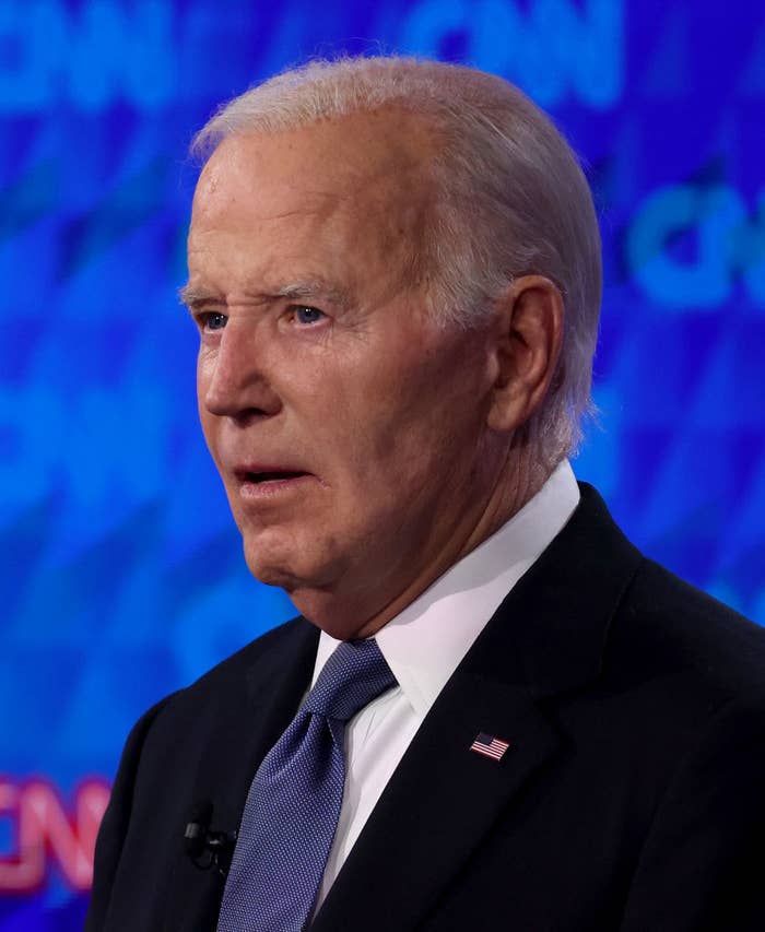 Joe Biden speaking during an event, wearing a suit and tie with an American flag pin on his lapel