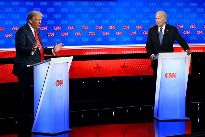 Donald Trump and Joe Biden on stage during a CNN debate, standing behind podiums. Trump is gesturing with his hands while Biden listens