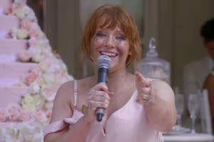 Bryce Dallas Howard gives a speech at a wedding reception, smiling and holding a microphone with a tiered floral wedding cake in the background