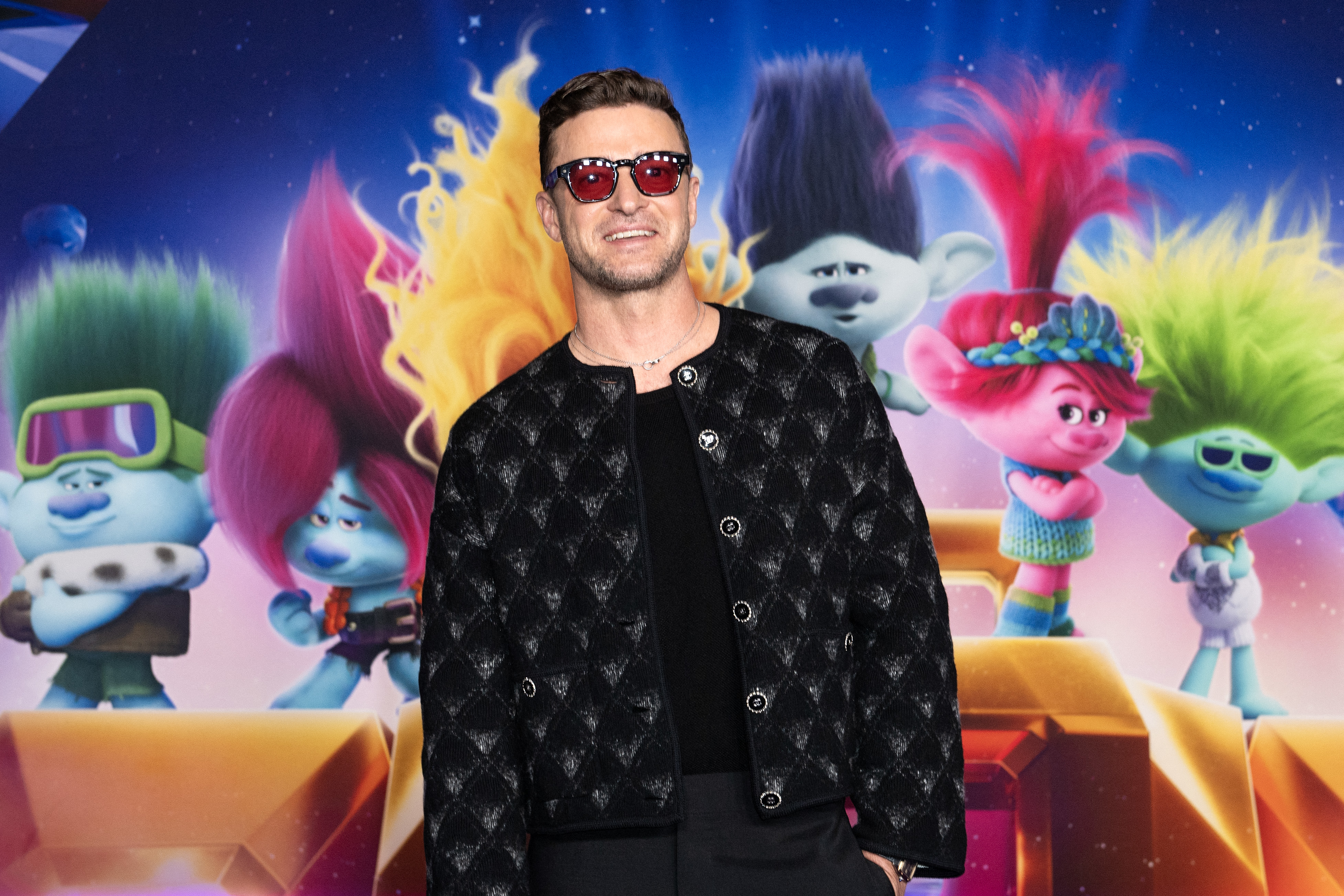 Justin Timberlake on the red carpet in front of a &quot;Trolls World Tour&quot; backdrop featuring animated characters. He wears a patterned jacket and sunglasses