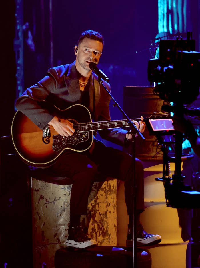 A man with short hair performs on stage, playing an acoustic guitar and singing into a microphone. Scene includes a camera recording him