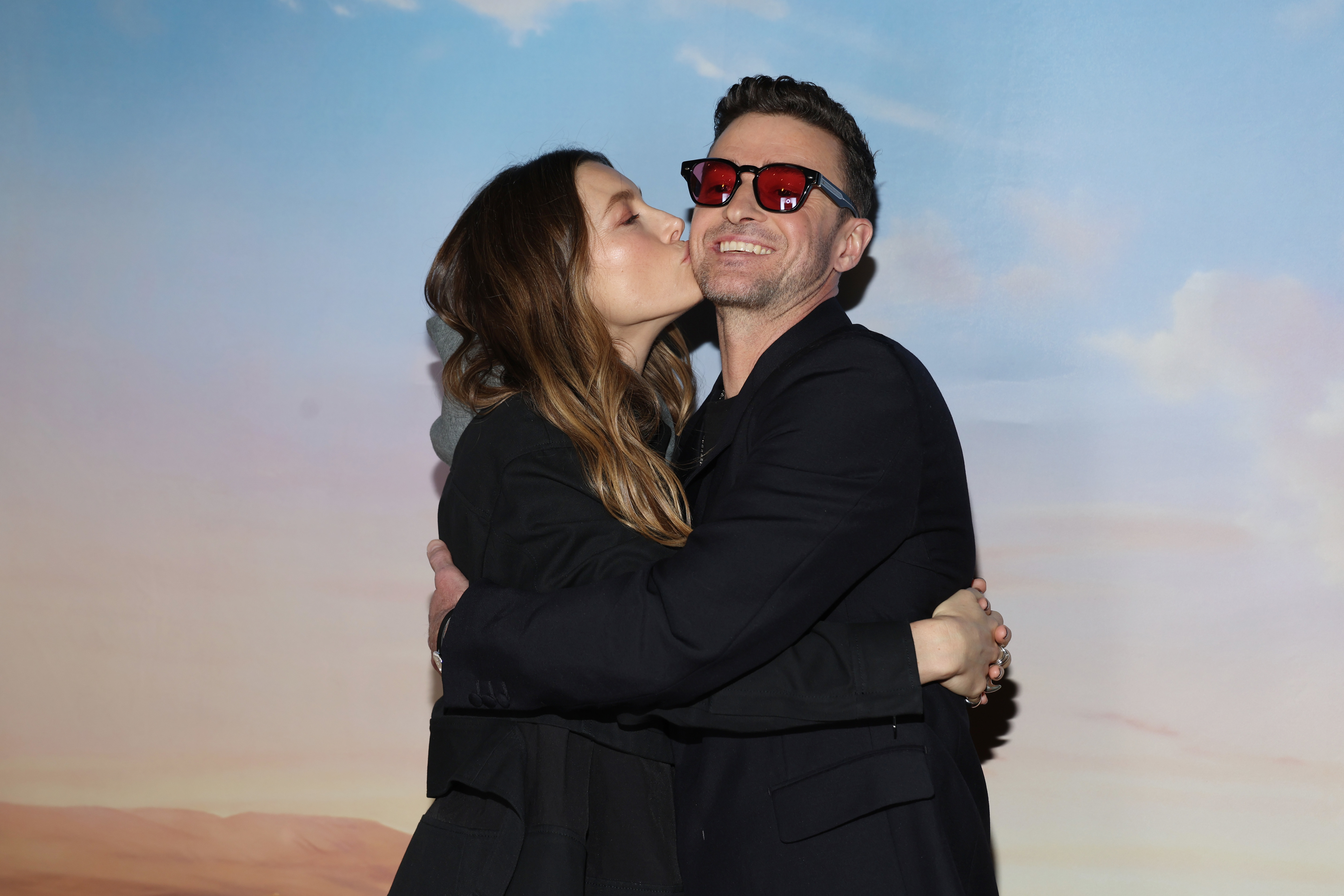 A woman wearing a black outfit kisses a smiling man&#x27;s cheek while they embrace. The man is wearing a black outfit and red sunglasses