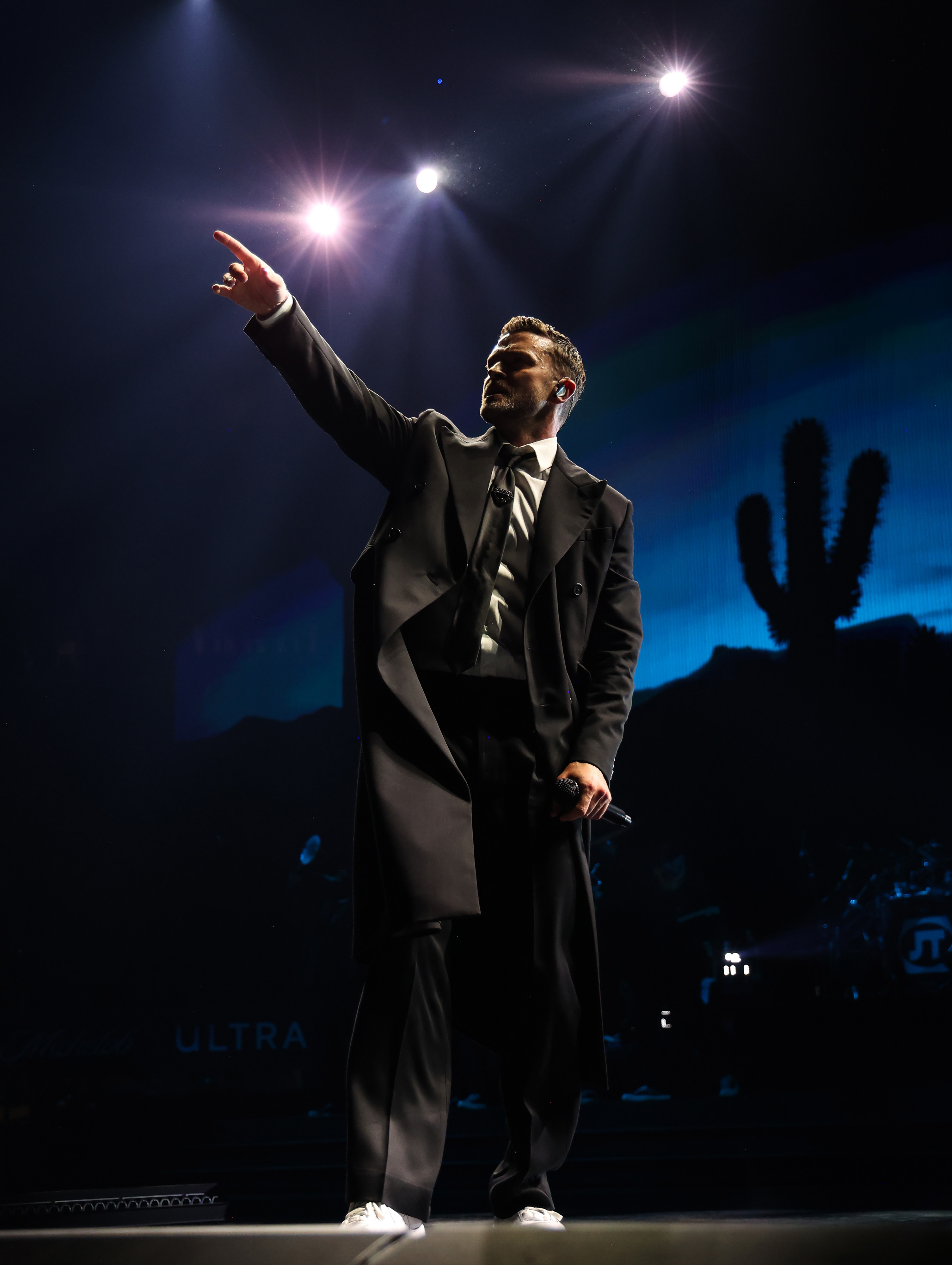 Justin Timberlake performs on stage in a suit with a long coat, pointing upward. A desert backdrop with a cactus is visible behind him
