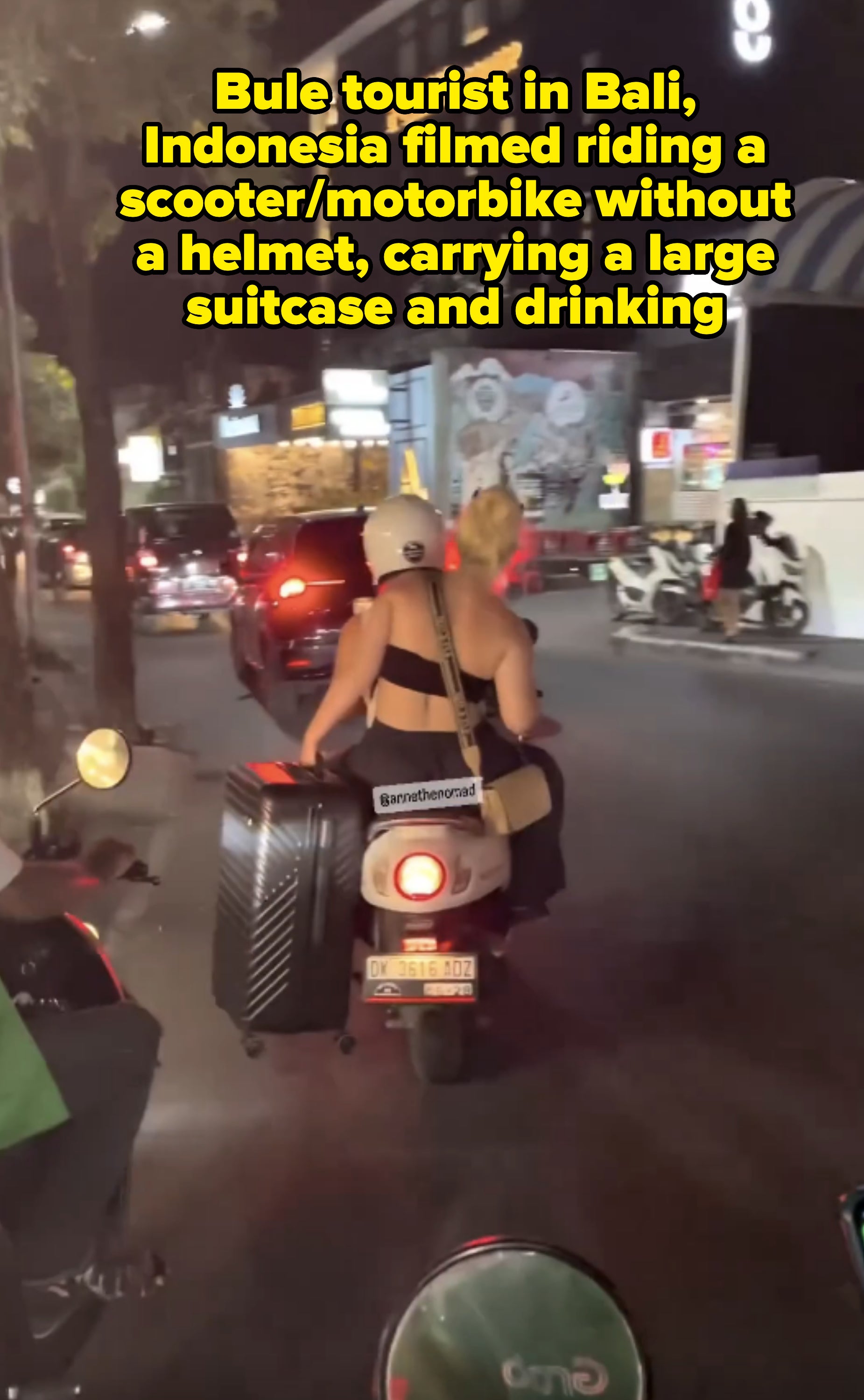 A woman on a motor scooter carries a large suitcase and a shoulder bag while riding through a busy city street at night