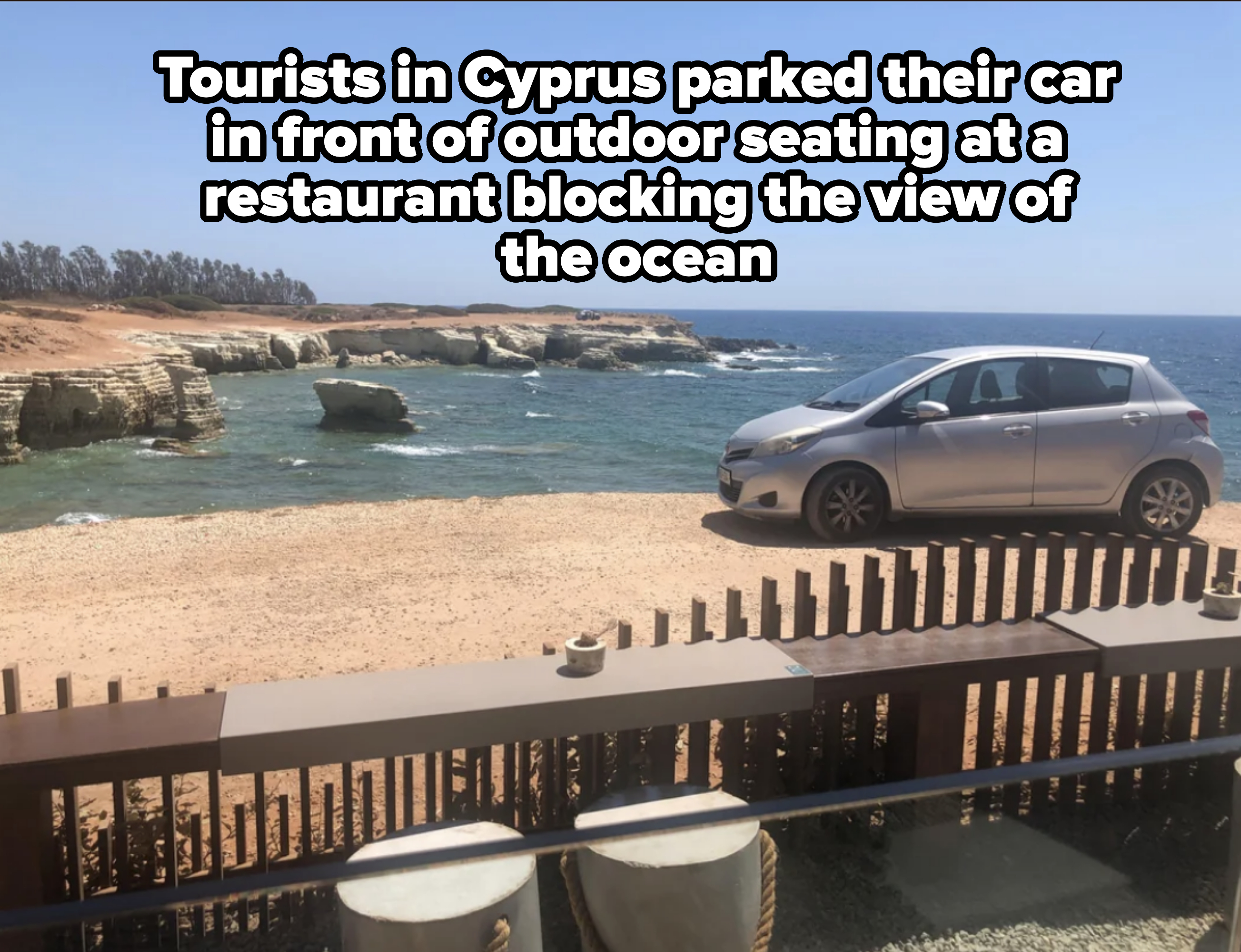 A silver car is parked on a sandy beach near rocky cliffs by a blue ocean under a clear sky. A wooden fence is in the foreground