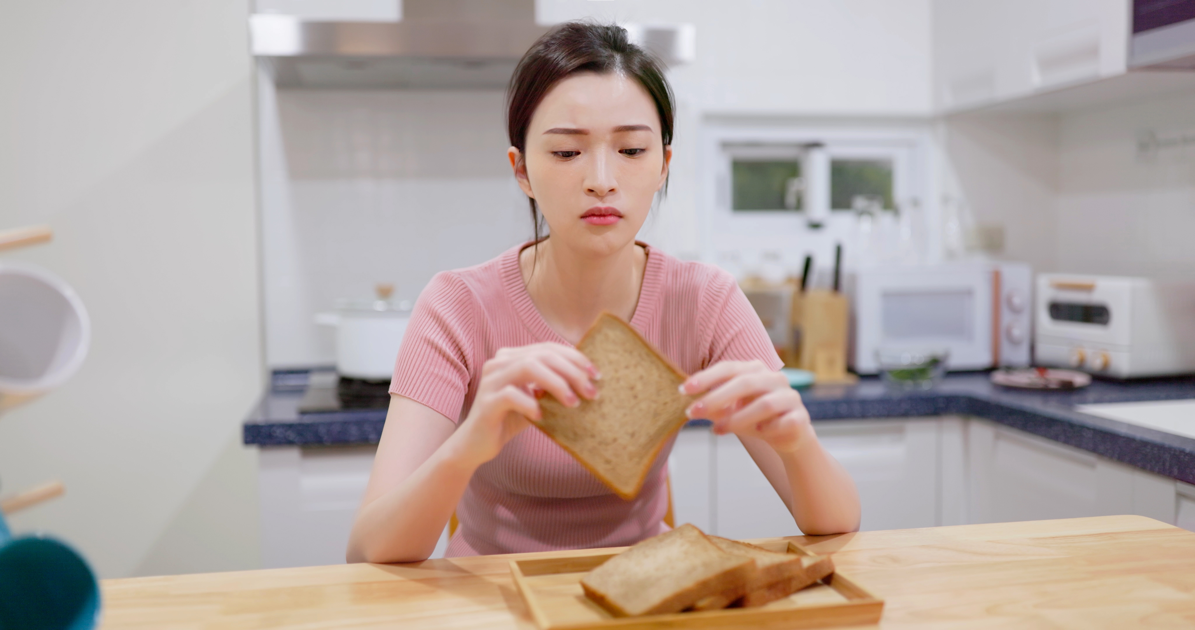 Woman in a kitchen looking thoughtfully at a slice of bread, with more slices on a plate in front of her