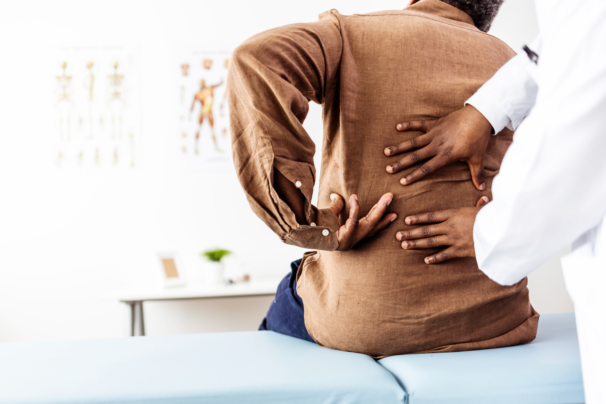 A patient with back pain, wearing a brown shirt, is being examined by a healthcare professional in a white coat, who is placing hands on the patient&#x27;s back