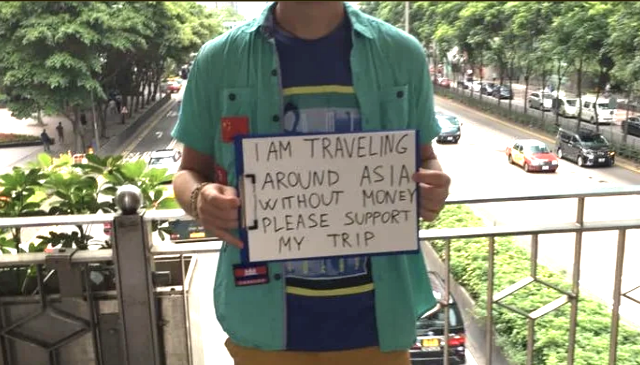 A person holding a sign that says, &quot;I am traveling around Asia without money. Please support my trip.&quot; The background shows a city street with cars