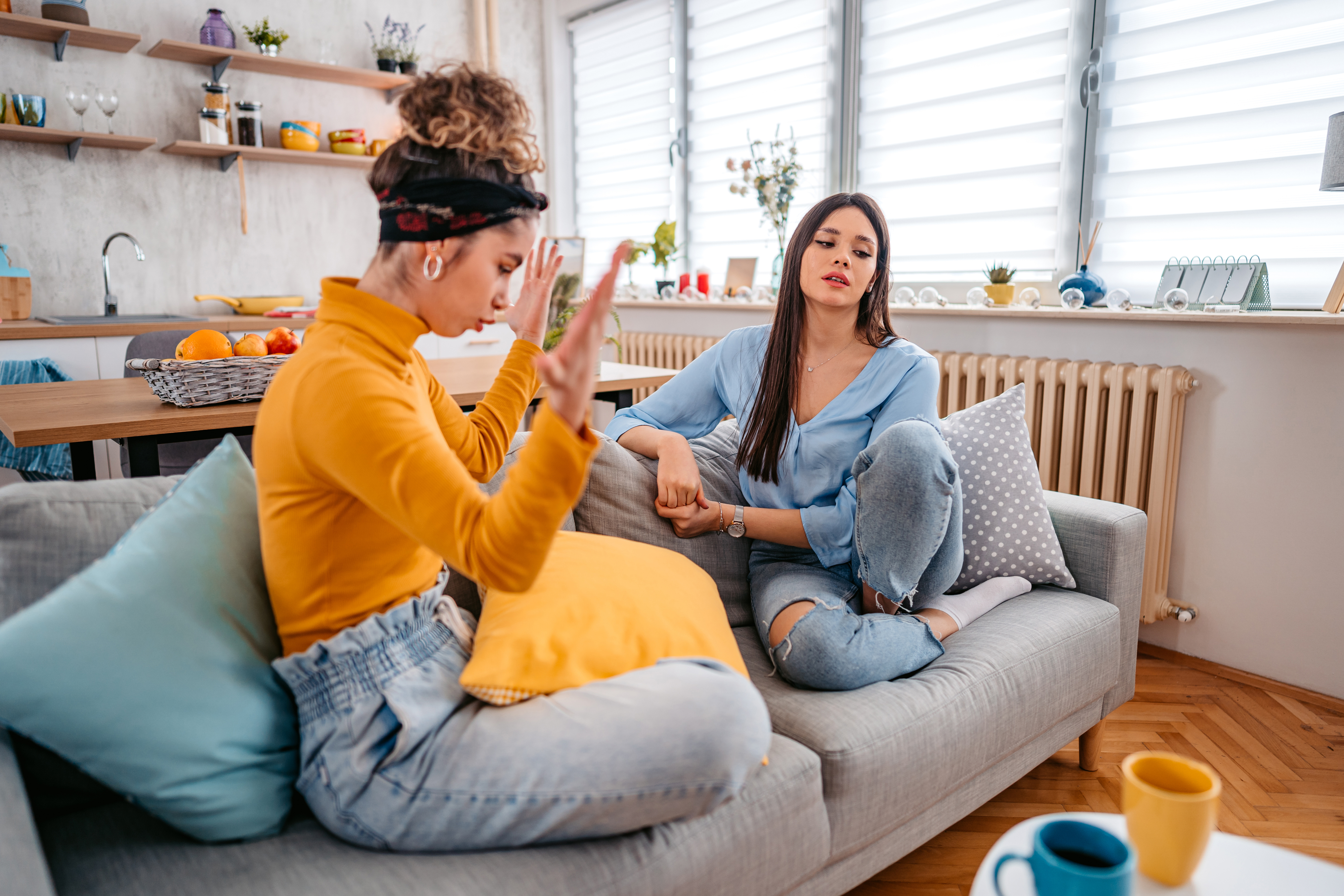 Two women sit on a couch in a casual setting, engaged in conversation. One woman gestures animatedly with her hands, the other listens attentively