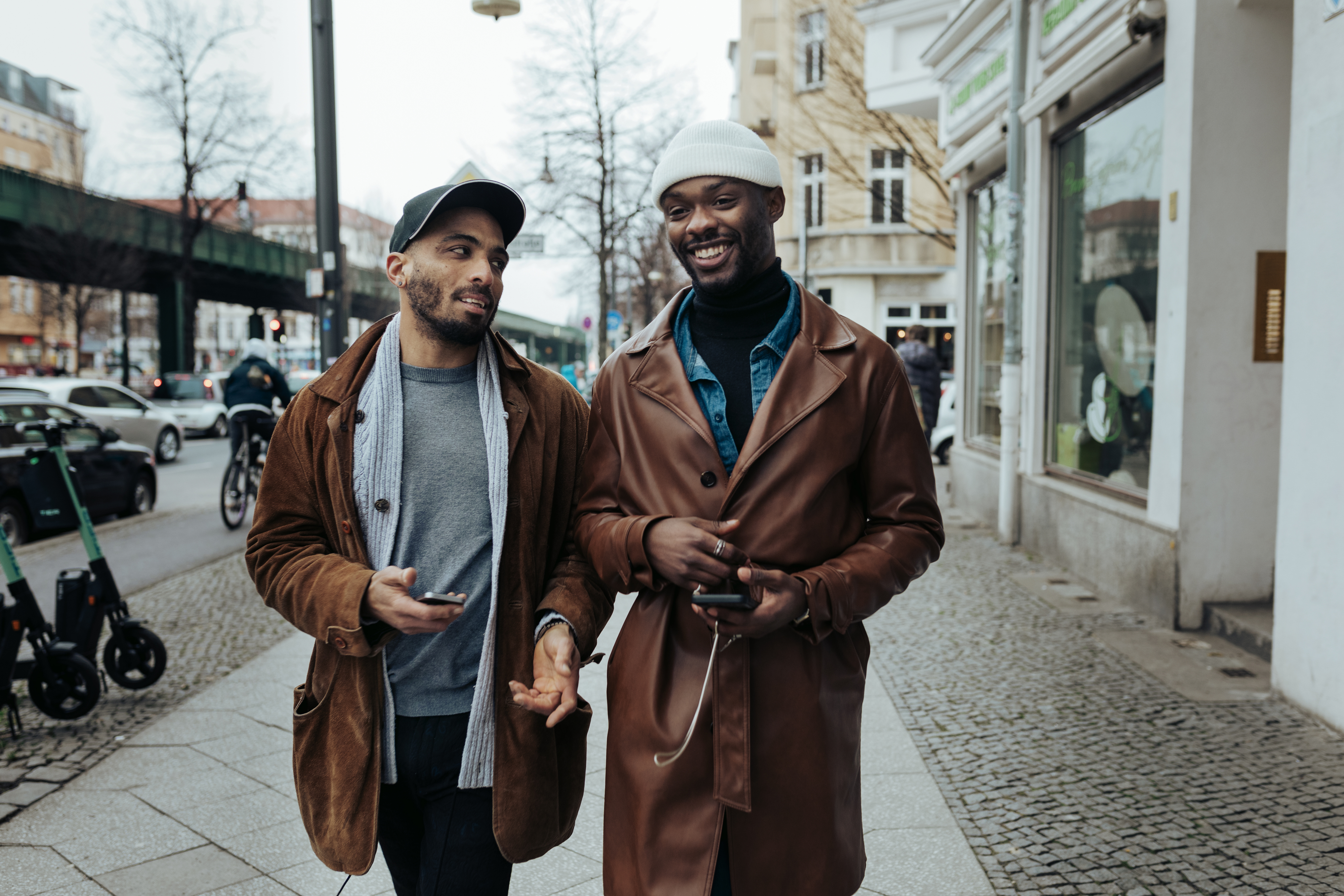 Two men, both wearing hats and brown coats, walk on a city sidewalk smiling and holding smartphones. They appear to be having a friendly conversation