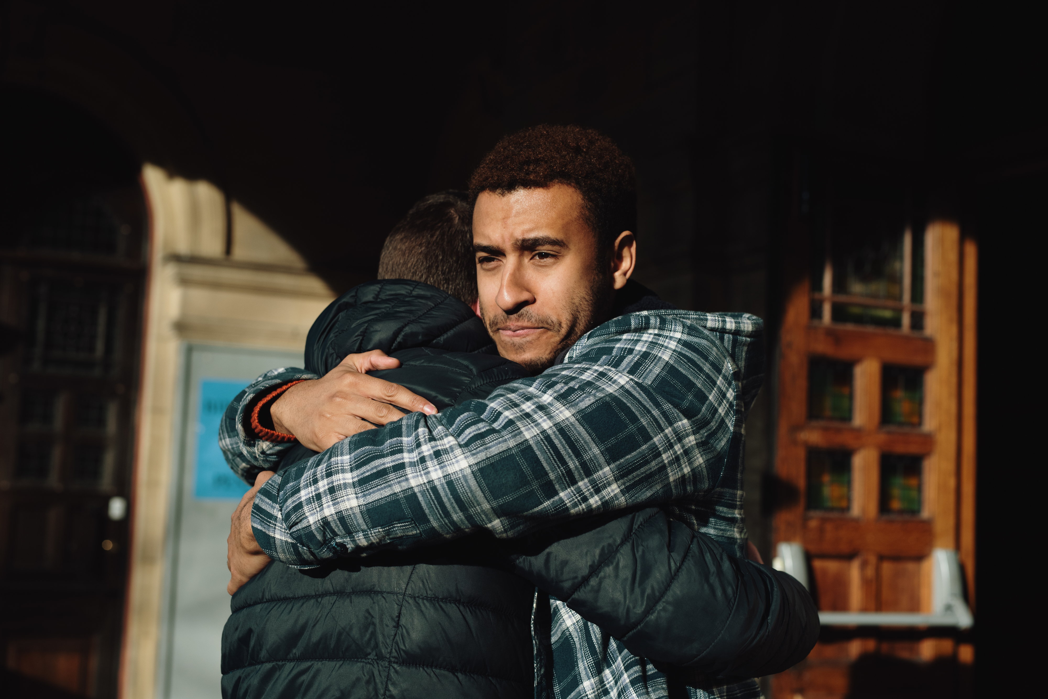 Two people are embracing in a warm hug outside a building, expressing affection and comfort. One person has curly hair and is wearing a plaid shirt