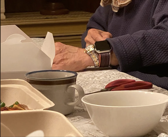 An elderly person&#x27;s hands are seen on a table, one hand wearing a smartwatch and a bracelet, a white takeout container, a mug, and food containers are nearby