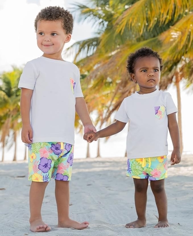 Two young children holding hands on a beach, wearing white T-shirts and colorful floral shorts. Palm trees in the background