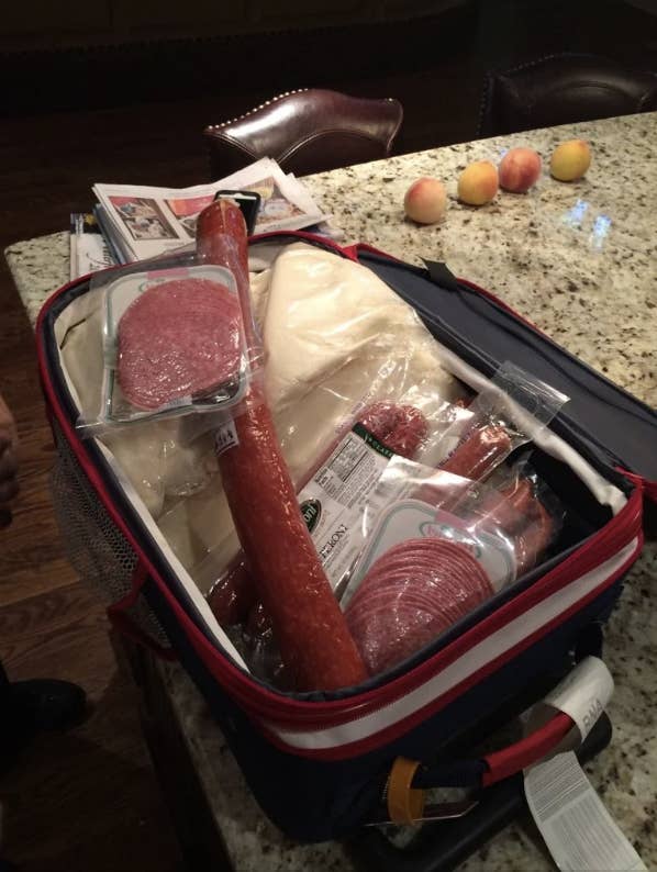 Open cooler on a kitchen counter filled with various meats, cheese slices, and a wrapped baguette. Peaches and newspapers are in the background