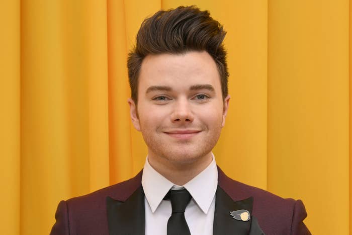 Chris Colfer smiles wearing a suit with a pin on the lapel, against a plain backdrop