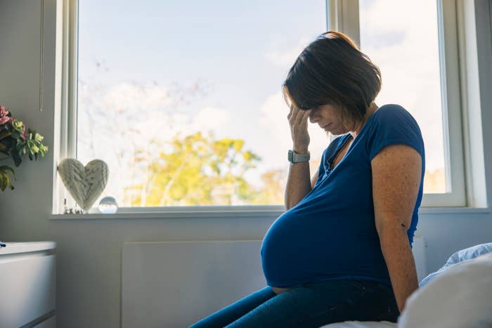 A pregnant woman sits on a bed, holding her head in apparent discomfort, near a window with daylight streaming in