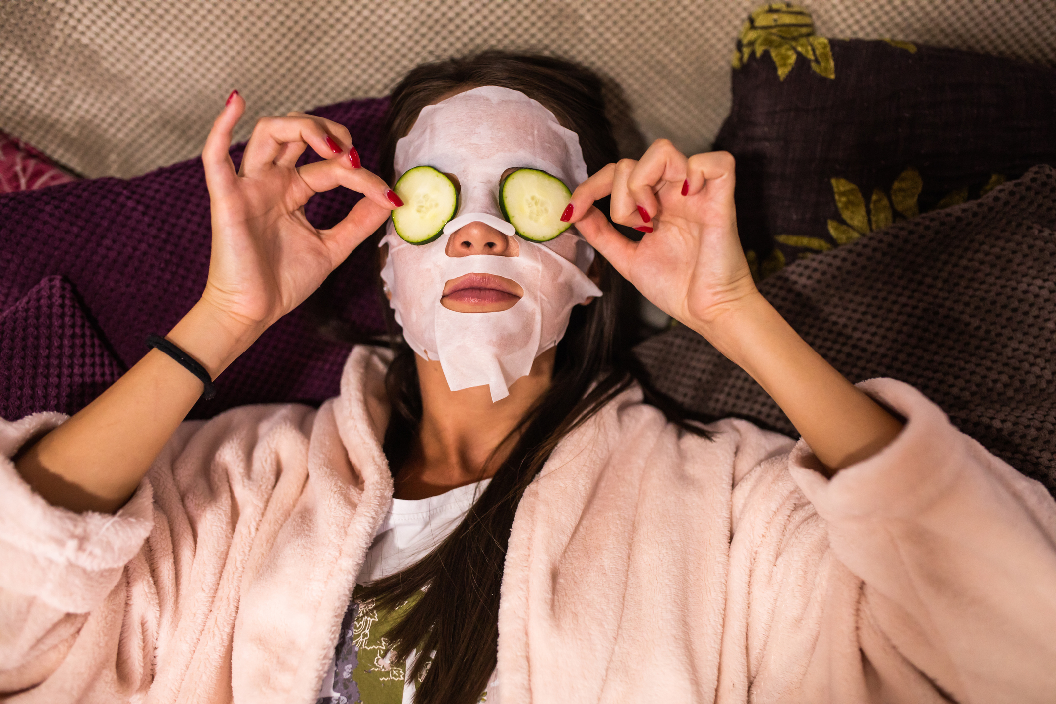 A person lies down wearing a sheet mask and holding cucumber slices over their eyes, enjoying a relaxing self-care routine