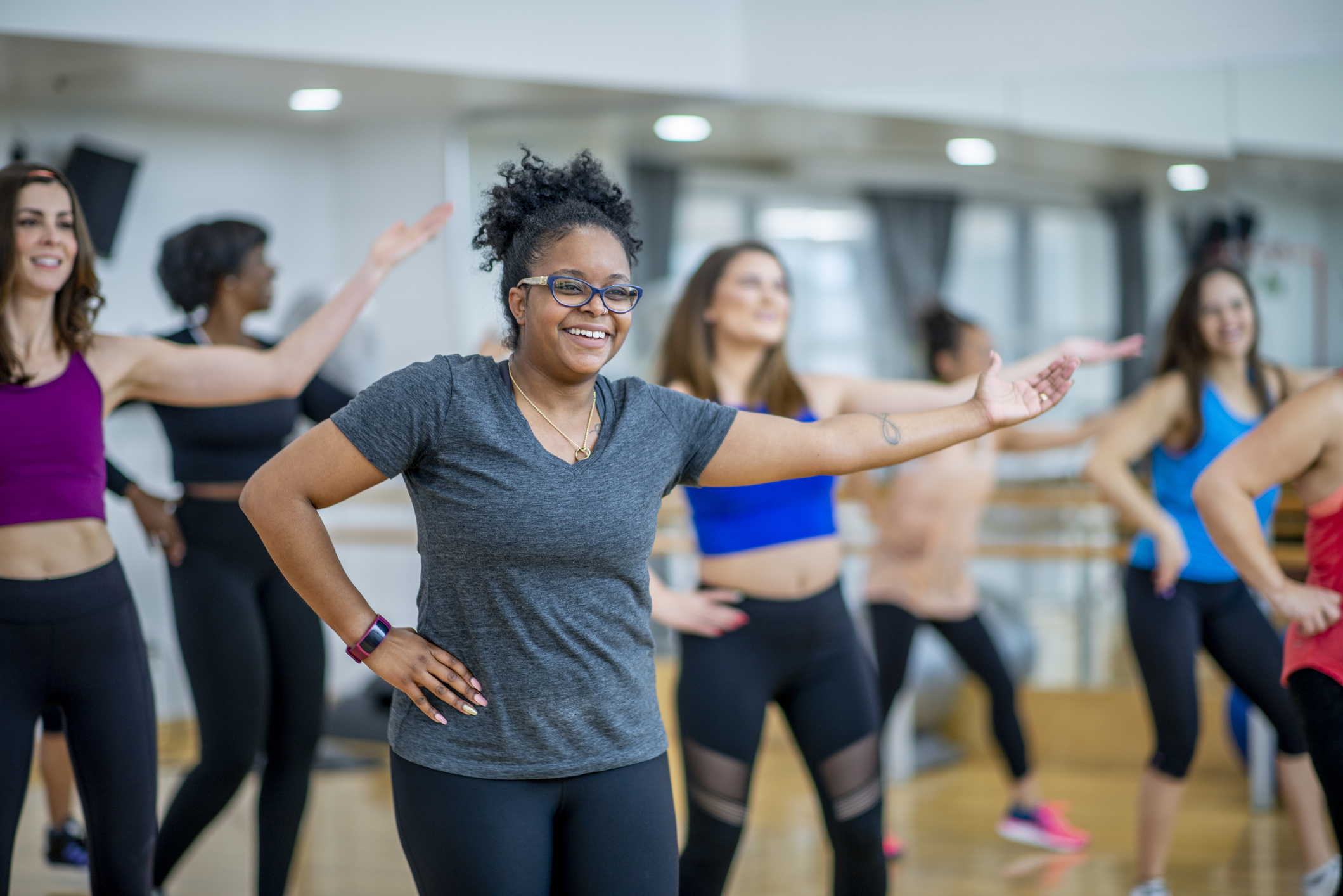 Group fitness class participants, including smiling woman in gray shirt and glasses, engage in an energetic workout session in a gym studio