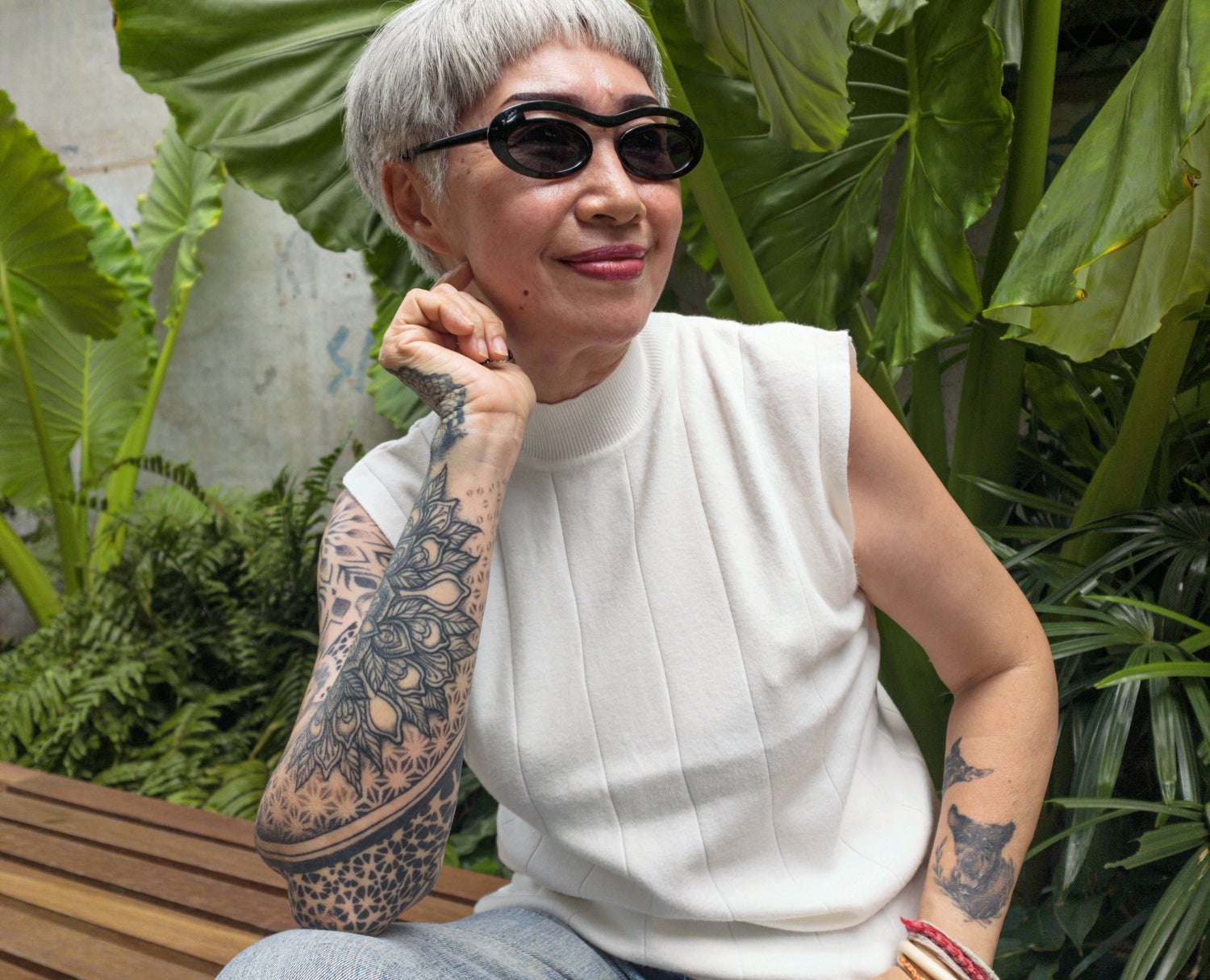 An elderly woman with tattoos smiles while sitting on a bench, wearing sunglasses, a sleeveless white top, and jeans, in a lush garden setting