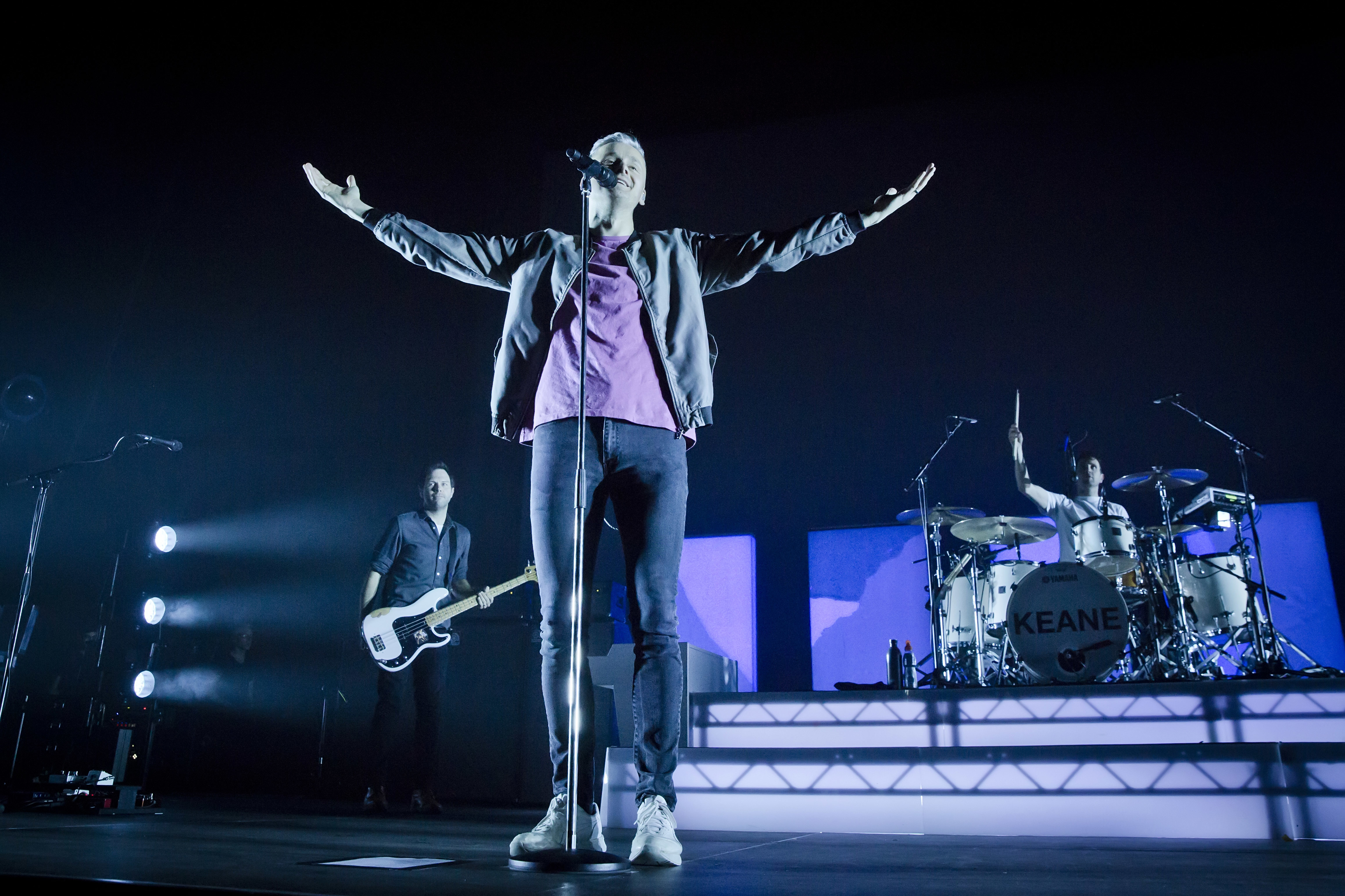 Keane performing on stage. The vocalist stands with arms outstretched while a guitarist and drummer play in the background