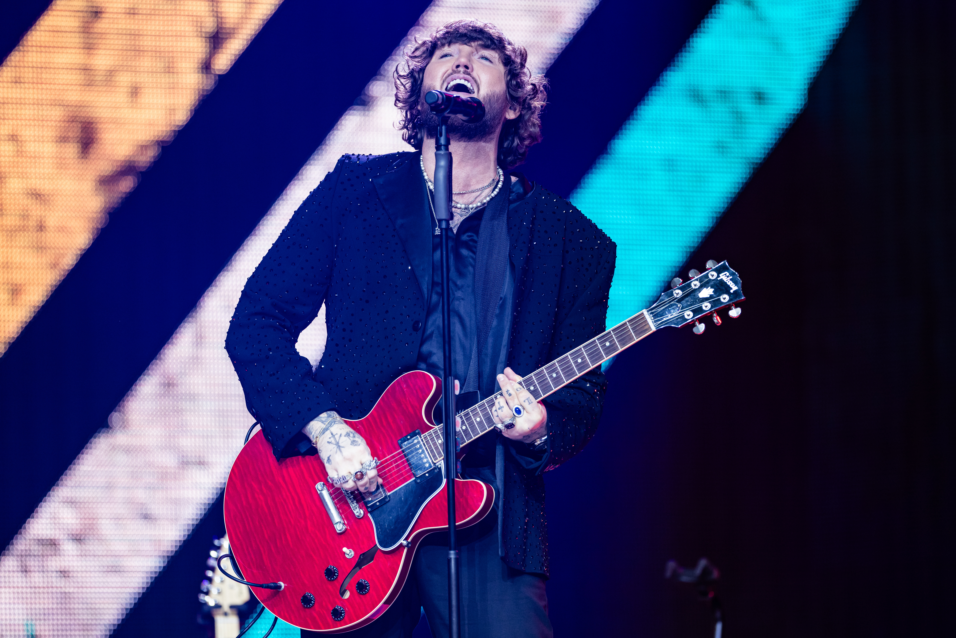 Person performing on stage with a bright red electric guitar, wearing a black outfit with sparkling details, and singing passionately into a microphone