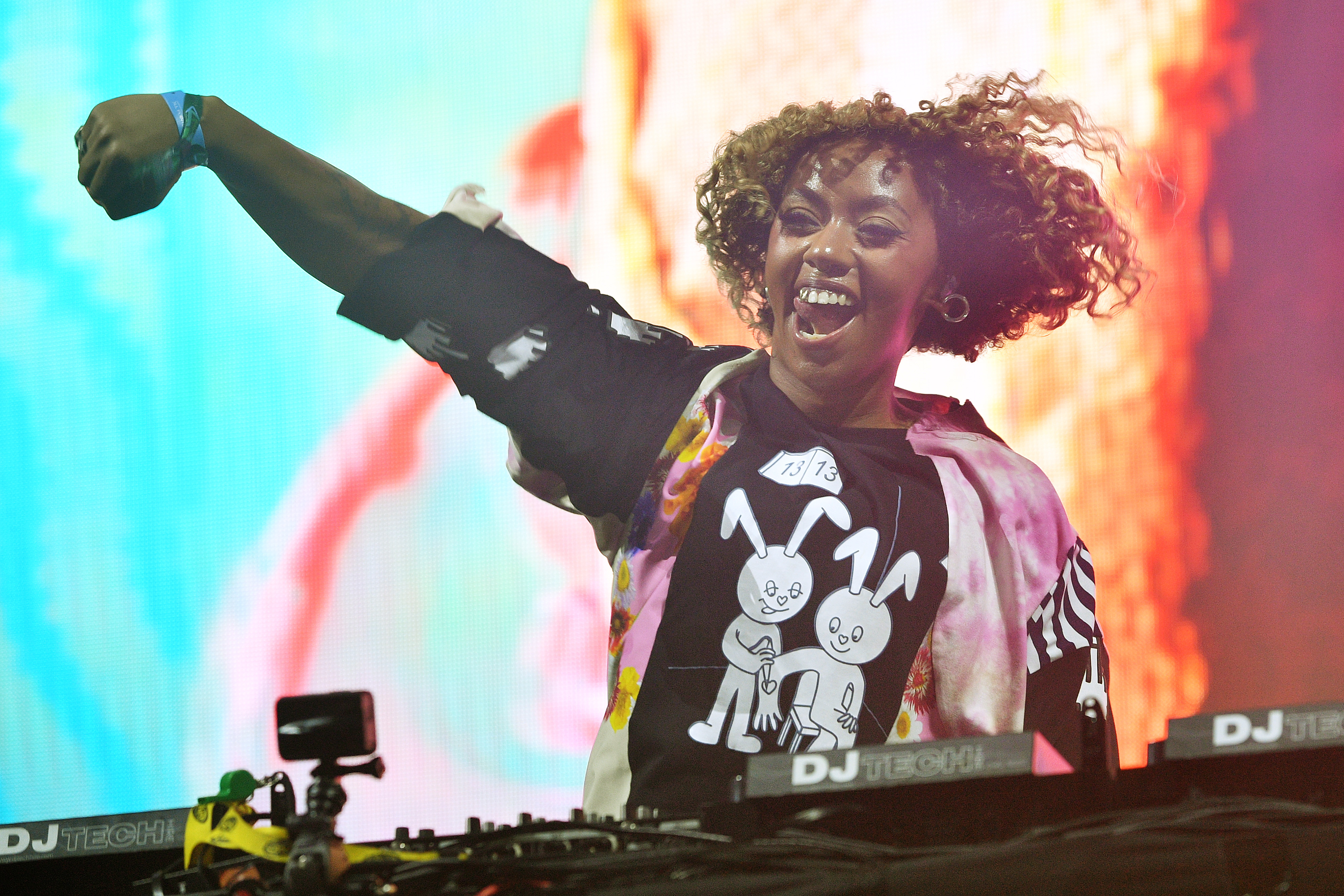 DJ LP Giobbi performs energetically, raising a fist, and wearing a graphic shirt with bunny illustrations, standing at a DJ booth