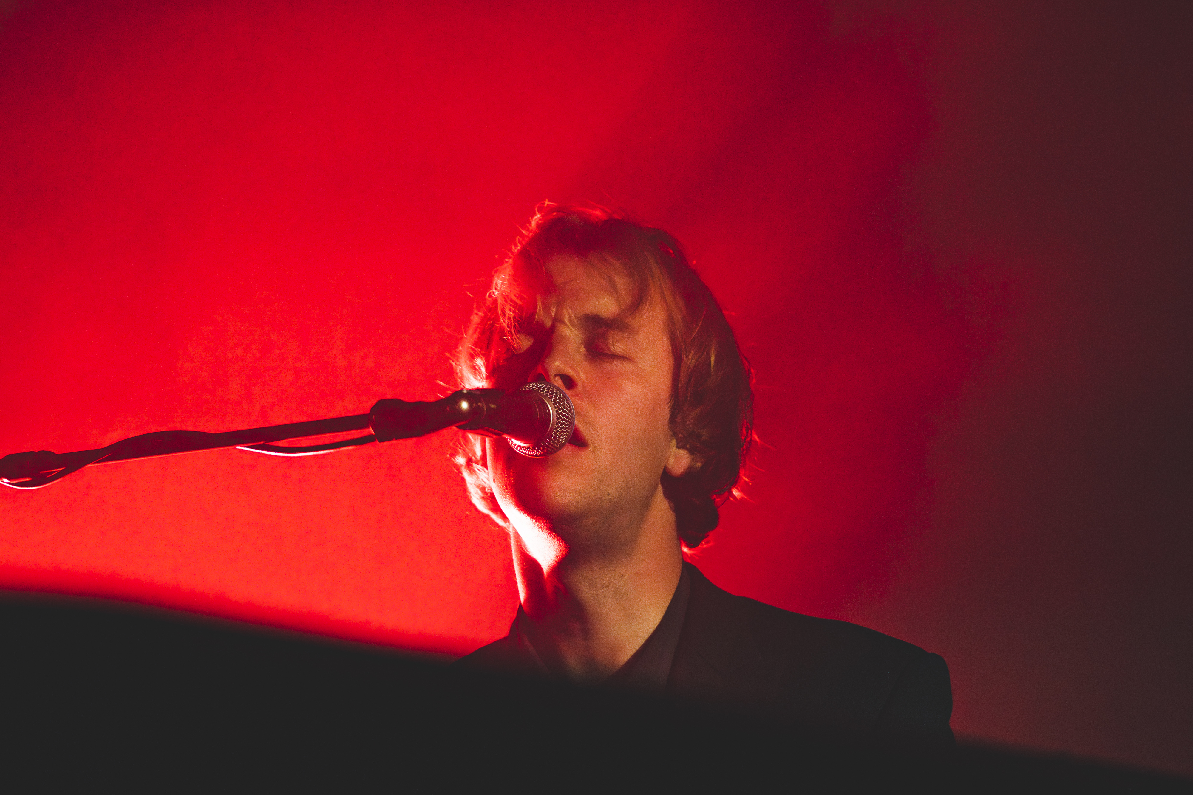 A person sings passionately into a microphone on stage, with a red light glowing in the background