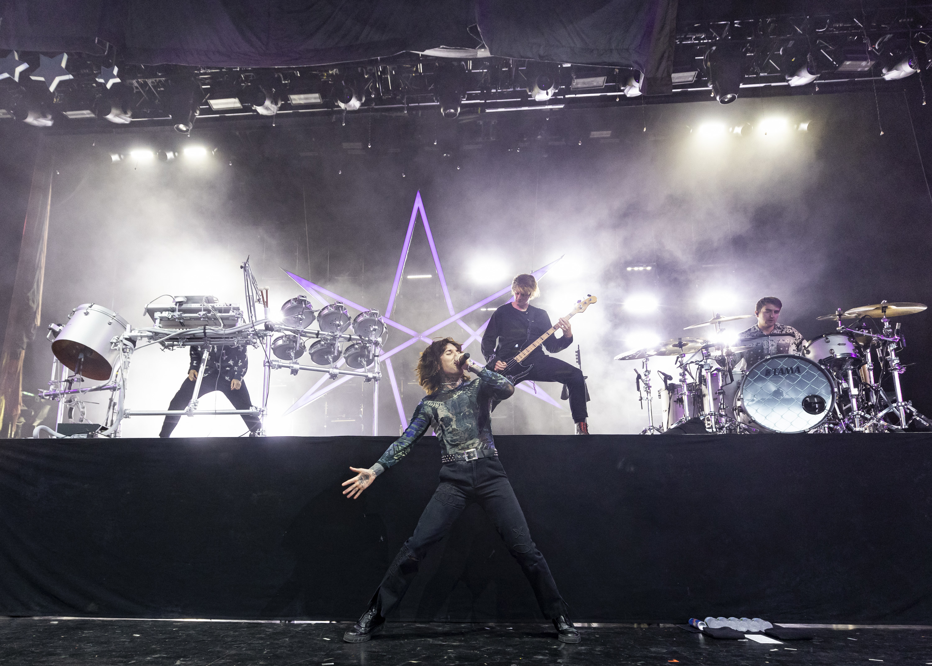 A band performs on stage with dramatic lighting. The lead singer passionately sings, while other members play drums and guitar in the background